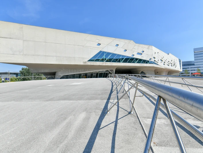 Modernes Betongebäude mit futuristischer Form und schrägen Glasflächen unter blauem Himmel.