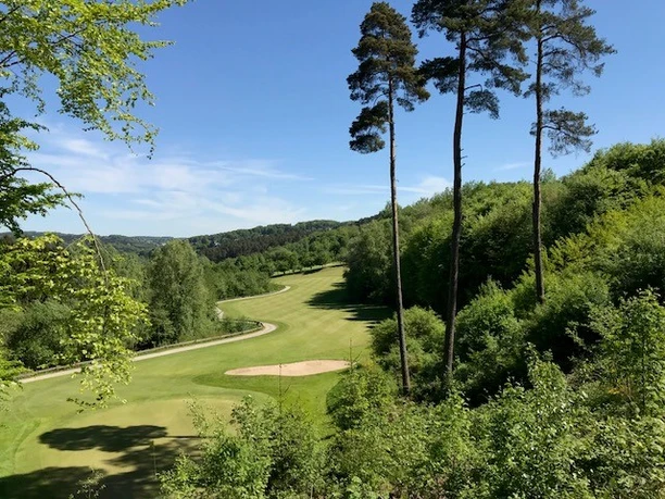 Golfclub Lüderich Grüner Golfplatz umgeben von dicht bewaldeten Hügeln unter klarem blauem Himmel, vereinzelt hohe Bäume.