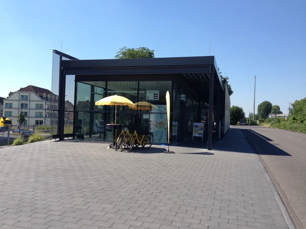Radstation Bergisch Gladbach Modernes Gebäude mit Glasfront, gelben Sonnenschirmen und Leihfahrrädern auf einer gepflasterten Fläche.