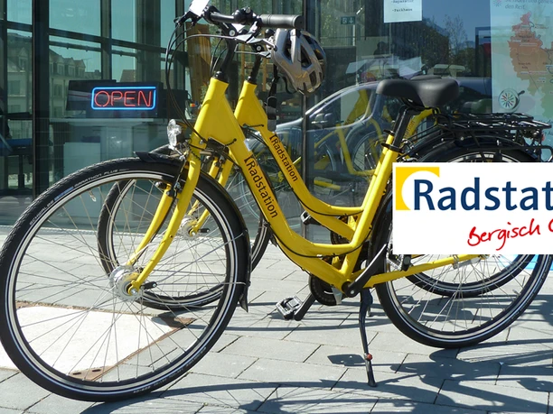 Radstation Bergisch Gladbach Zwei gelbe Leihräder vor einer Glastür mit "OPEN"-Schild und Radstation Bergisch Gladbach Logo.