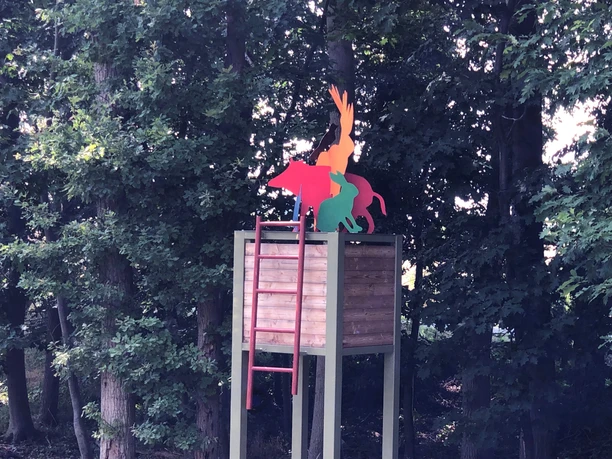Hochsitz Ein Hochsitz flankiert von Bäumen mit kunterbunten Tierfiguren auf der Balustrade in einem Wald.