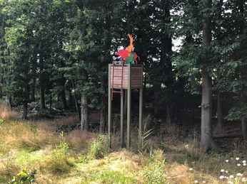 Hochsitz Ein Hochsitz umgeben von Bäumen und hohem Gras, mit bunten Figuren auf dem Dach.