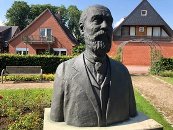 Bronzestatue eines bärtigen Mannes im Anzug vor roten Backsteinhäusern und gepflegtem Garten.