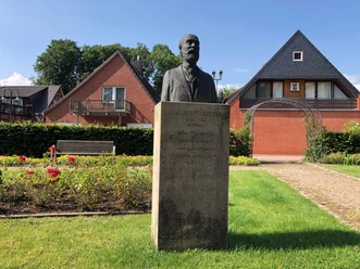 Skulptur von Adelbert Hotzen steht vor einem Ziegelhaus, umgeben von gepflegten Grünflächen.