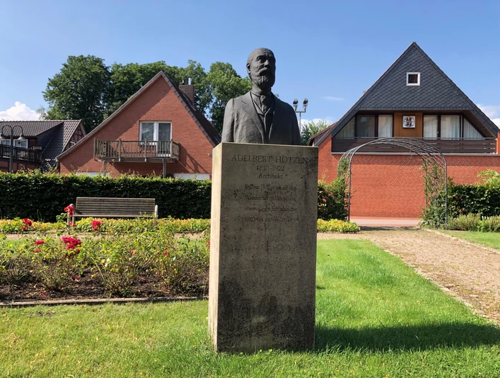 Adelbert Hotzen Skulptur von Adelbert Hotzen steht vor einem Ziegelhaus, umgeben von gepflegten Grünflächen.