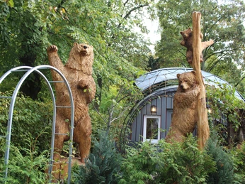 Zwei lebensgroße hölzerne Bärenskulpturen stehen in einem üppig grünen Garten vor einem modernen Gebäude.