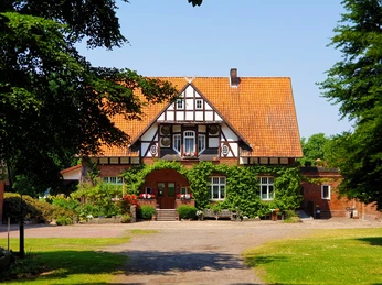 Ein historisches Fachwerkhaus mit rotem Ziegeldach, umgeben von grüner Landschaft und einem gepflasterten Weg.
