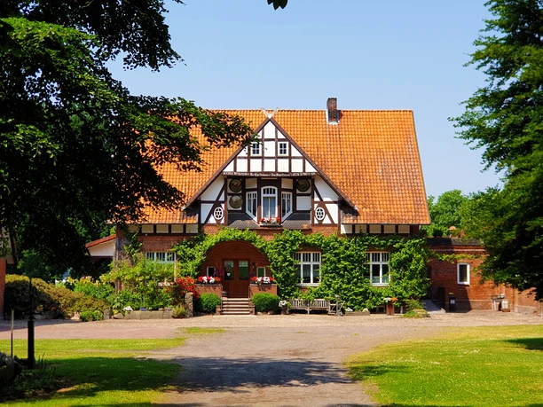 Ein historisches Fachwerkhaus mit rotem Ziegeldach, umgeben von grüner Landschaft und einem gepflasterten Weg.