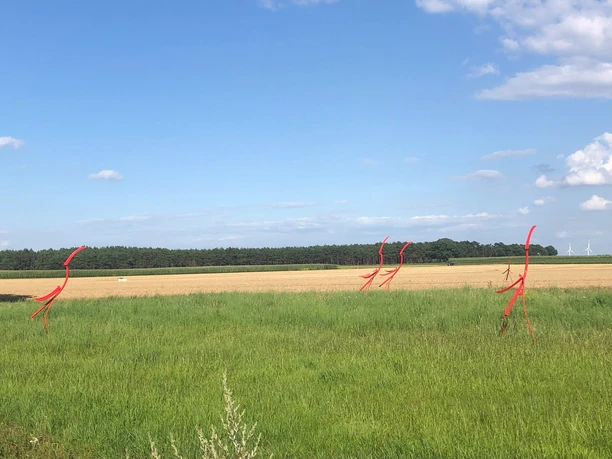 Rote, abstrakte Stabfiguren aus Metall stehen auf einer grünen Wiese unter einem klaren blauen Himmel.