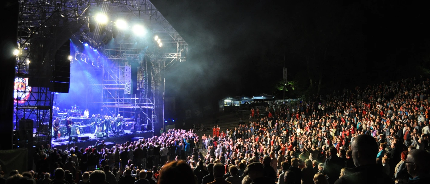 Concert on an illuminated open-air stage, surrounded by a large crowd at night.