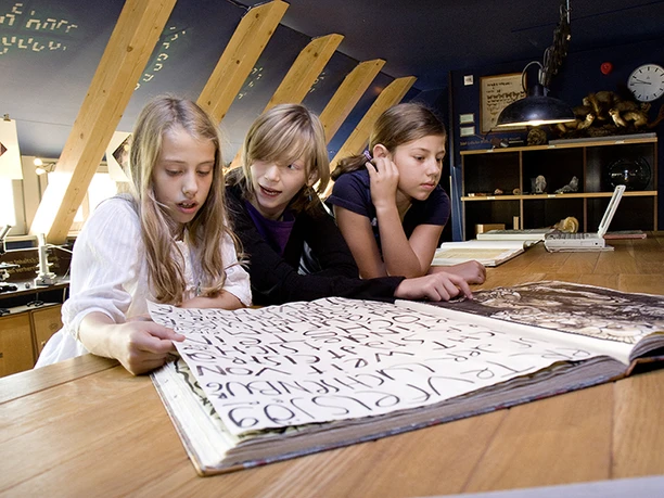 Three children look at a large open book in a museum room.