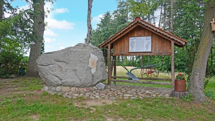 Mitte des Emslandes Großer Findling mit Inschrift und Infotafel unter Holzunterstand im Wald, daneben ein alter Pflug.