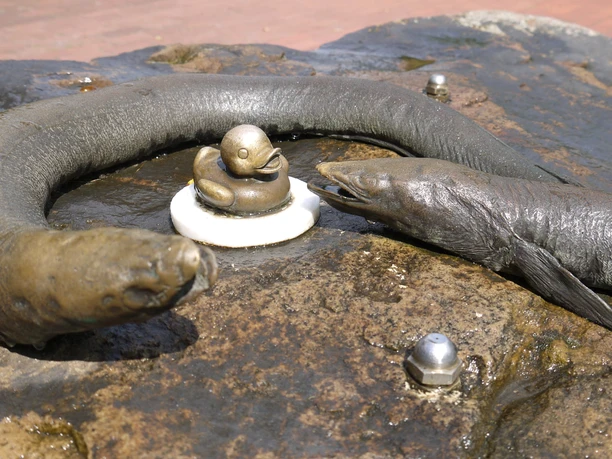 Aalisa-Aali-min.jpg Bronzefiguren von zwei Aalen umrahmen eine kleine Ente auf einem nassen Steinbrunnen in Bad Zwischenahn.