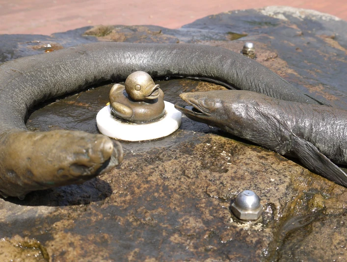 Aalisa-Aali-min.jpg Bronzefiguren von zwei Aalen umrahmen eine kleine Ente auf einem nassen Steinbrunnen in Bad Zwischenahn.