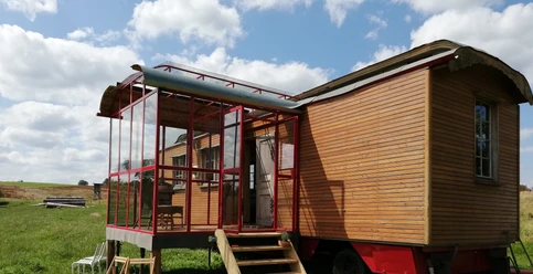 Ein hölzerner Zirkuswagen mit Glasveranda steht auf einer Wiese unter blauem Himmel mit Schäfchenwolken.