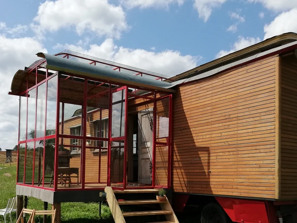 Außenansicht Ein hölzerner Zirkuswagen mit Glasveranda steht auf einer Wiese unter blauem Himmel mit Schäfchenwolken.