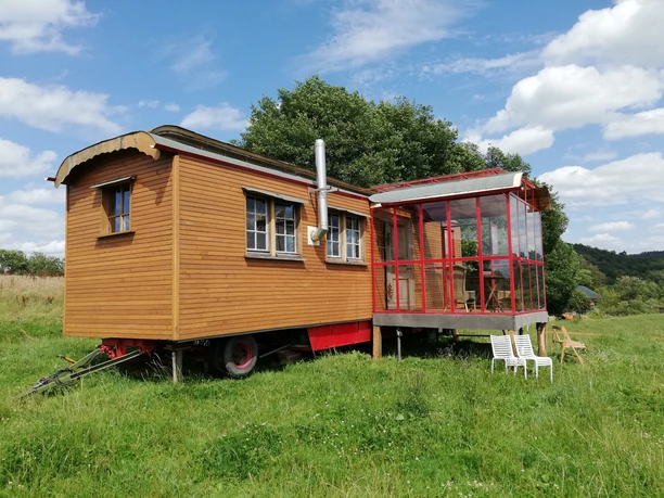 Außenansicht 2 Ein hölzerner Zirkuswagen mit verglastem Anbau steht auf einer grünen Wiese unter blauem Himmel.