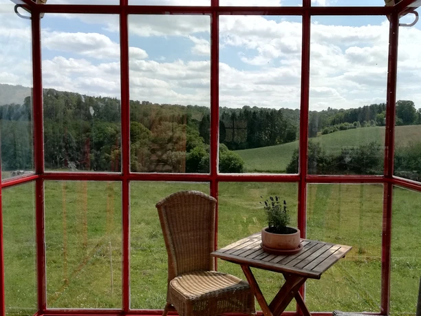 Aussicht Ein lichtdurchfluteter Wintergarten mit roten Rahmen, Holzfußboden, Blick auf grüne Hügel und Himmel.