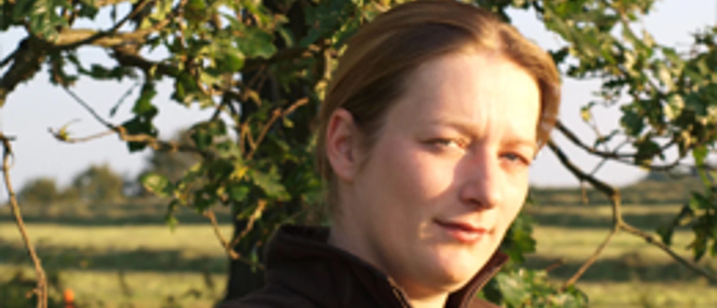 Bild Silke Junick.jpg Eine Frau steht vor einem Baum in einer ländlichen Umgebung, Sonnenlicht fällt auf ihr Gesicht.