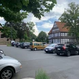 Auf einem Parkplatz stehen mehrere Autos, im Hintergrund befindet sich ein Fachwerkhaus und grüne Bäume.