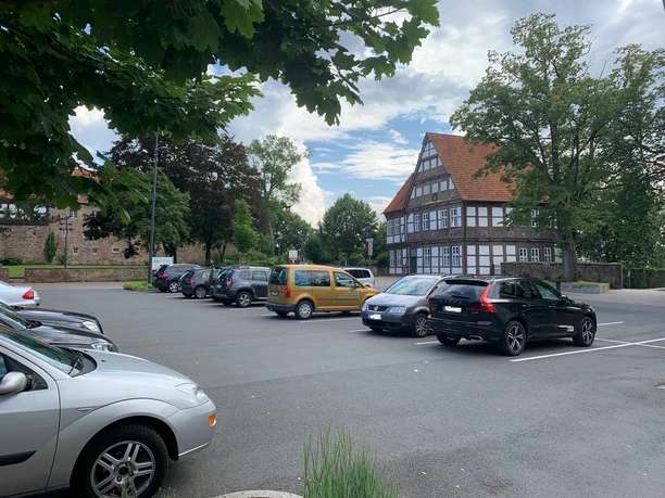 Auf einem Parkplatz stehen mehrere Autos, im Hintergrund befindet sich ein Fachwerkhaus und grüne Bäume.