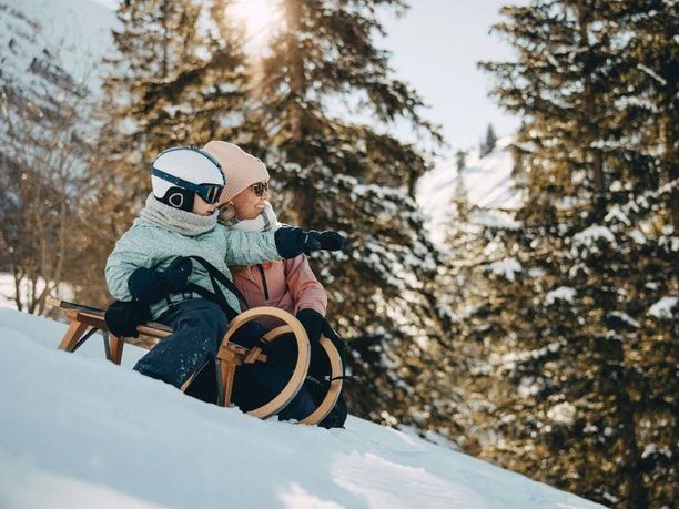 Snow-covered landscapes invite you to sledding fun Child sits on a wooden sled and points forward with his hand, mother kneels next to him