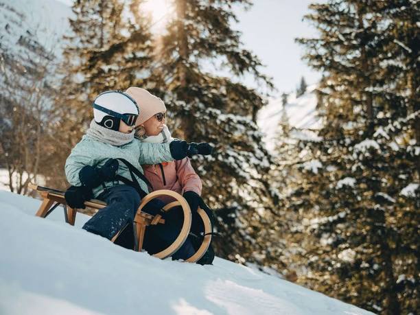 Les paysages enneigés invitent à faire de la luge L'enfant est assis sur une luge en bois et pointe la main vers l'avant, sa mère est agenouillée à côté de lui.