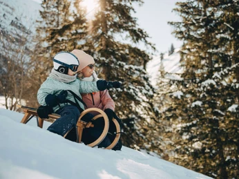 Verschneite Landschaften laden zum Schlittelplausch ein Kind sitzt auf einem Holzschlitten und zeigt mit der Hand nach vorne, die Mutter kniet danebenChild sits on a wooden sled and points forward with his hand, mother kneels next to himL'enfant est assis sur une luge en bois et pointe la main vers l'avant, sa mère est agenouillée à côté de lui.