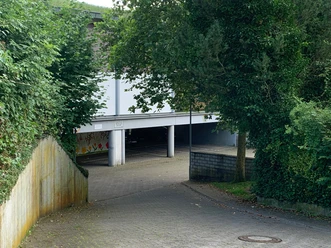 Parkpalette "Am Weinberg" Parkhaus bei Weinreben, umgeben von Bäumen, mit Zufahrtsrampe und Graffitis, teils im Schatten.