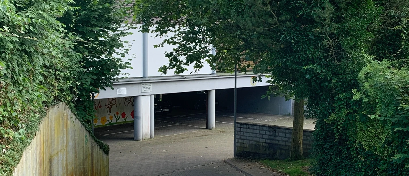 Parkhaus bei Weinreben, umgeben von Bäumen, mit Zufahrtsrampe und Graffitis, teils im Schatten.