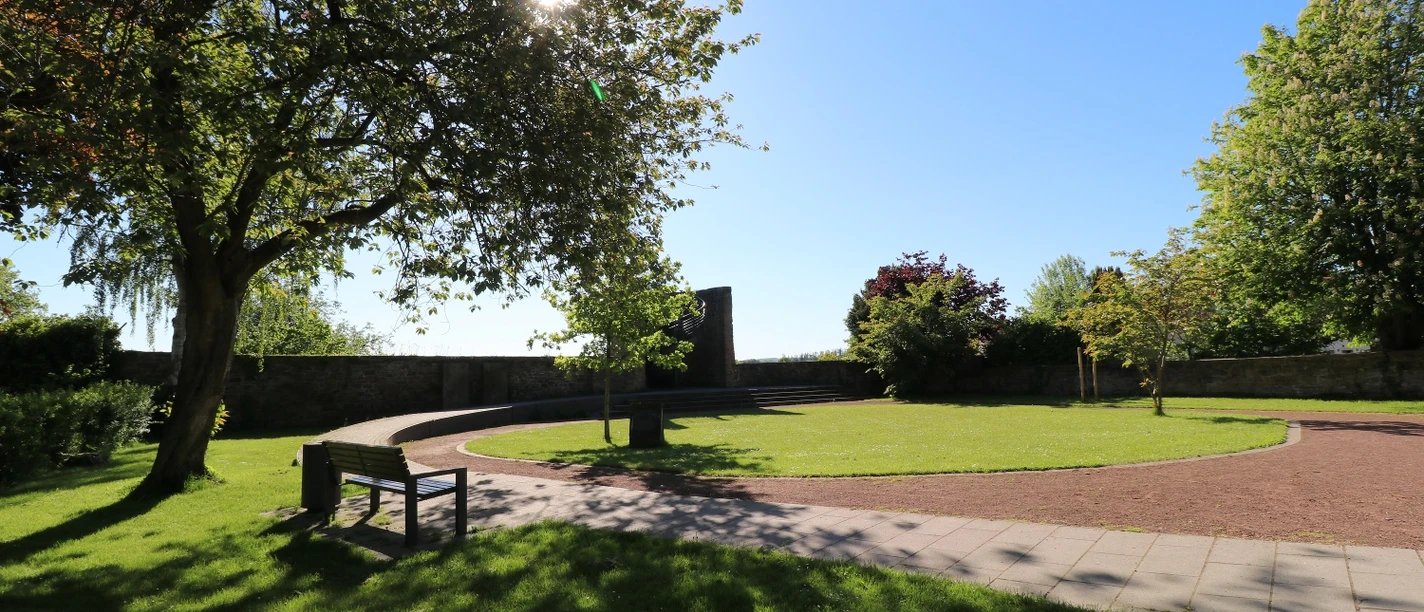 Stadtpark im Sonnenlicht mit geschwungenen Wegen, Bänken und schattenspendenden Bäumen.