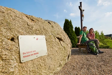 Ubben Krüß Zwei Wandernde sitzen auf einer Bank neben einem Holzkreuz und einem Findling mit Pilgerwegschild.