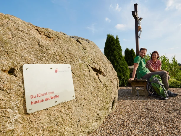 Ubben Krüß Zwei Wandernde sitzen auf einer Bank neben einem Holzkreuz und einem Findling mit Pilgerwegschild.