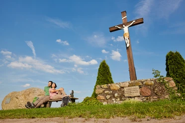 Ubben Krüß Ein Paar ruht auf einer Bank neben einem großen Holzkreuz unter blauem Himmel im Naturpark Hümmling.