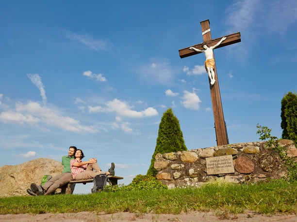 Ubben Krüß Ein Paar ruht auf einer Bank neben einem großen Holzkreuz unter blauem Himmel im Naturpark Hümmling.