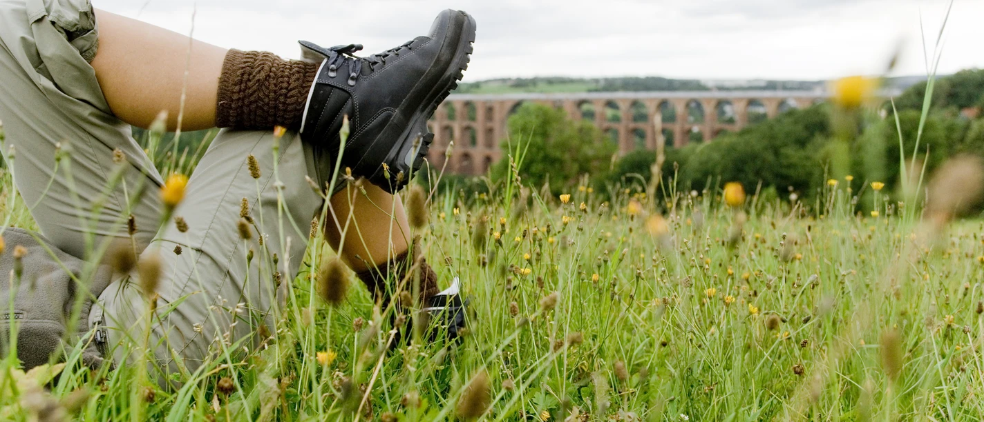 rene-gaens-7930.jpg Person in Wanderschuhen liegt im Gras, im Hintergrund ein historisches Viadukt.