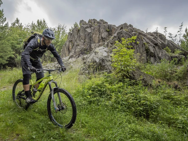 Ein Mountainbiker fährt an einem felsigen Hügel in einer grünen, bewaldeten Umgebung vorbei.
