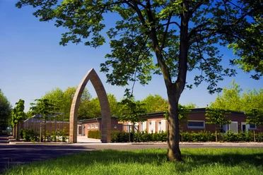 Kloster Esterwegen Moderner Torbogen aus Holz am Eingang des Klosters Esterwegen mit Gebäuden und Bäumen im Hintergrund.