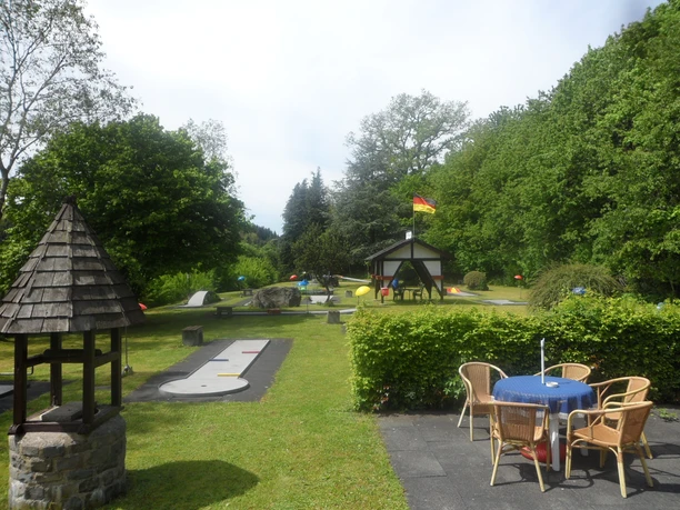 Minigolfplatz in Reichshof-Eckenhagen Minigolfplatz im Grünen mit Holztisch, Stühlen und Schirm, umgeben von Bäumen und Sträuchern.
