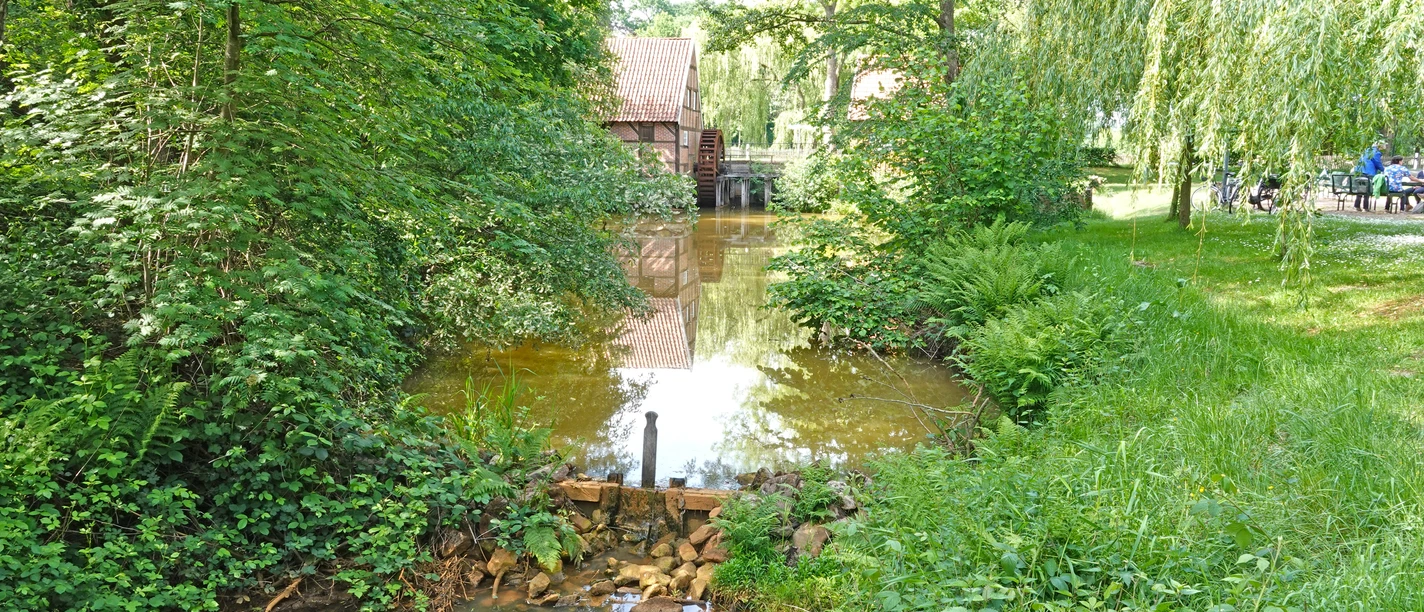 Wassermühle Bruneforth ©Emsland Tourismus Alte Wassermühle mit hölzernem Mühlrad an einem Bachlauf, umgeben von Bäumen und dichtem Grün
