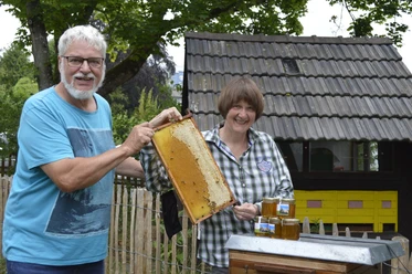 Bienen Museumshonig Ein älteres Paar präsentiert stolz eine mit Honig gefüllte Bienenwabe, im Hintergrund ein Bienenhaus.