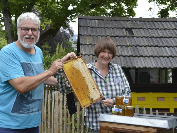 Bienen Museumshonig Ein älteres Paar präsentiert stolz eine mit Honig gefüllte Bienenwabe, im Hintergrund ein Bienenhaus.