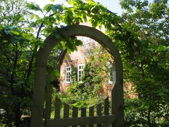 OLYMPUS DIGITAL CAMERA Ein großes Holztor führt durch saftig grünes Laub zu einem traditionellen Backsteingebäude.