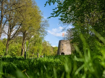 Kumsttonne Oerlinghausen "Frühsommeransichten" Alte Windmühle aus Stein in grünem Park mit blauem Himmel und vereinzelten Wolken im Hintergrund.