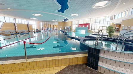 Ein modernes Schwimmbad bietet ein großes Becken mit klar abgegrenzten Schwimmbahnen und eine sanfte, angenehme Beleuchtung, die durch Fenster Tageslicht hereinlässt. Das Design ist einladend und barrierefrei gestaltet.