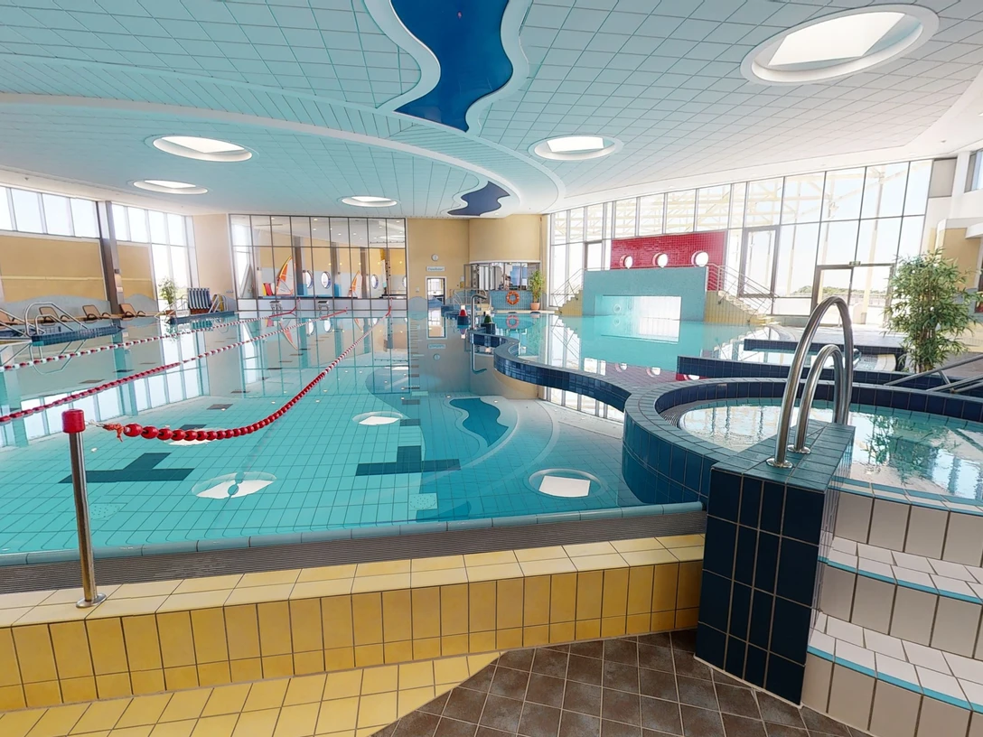 Ein modernes Schwimmbad bietet ein großes Becken mit klar abgegrenzten Schwimmbahnen und eine sanfte, angenehme Beleuchtung, die durch Fenster Tageslicht hereinlässt. Das Design ist einladend und barrierefrei gestaltet.