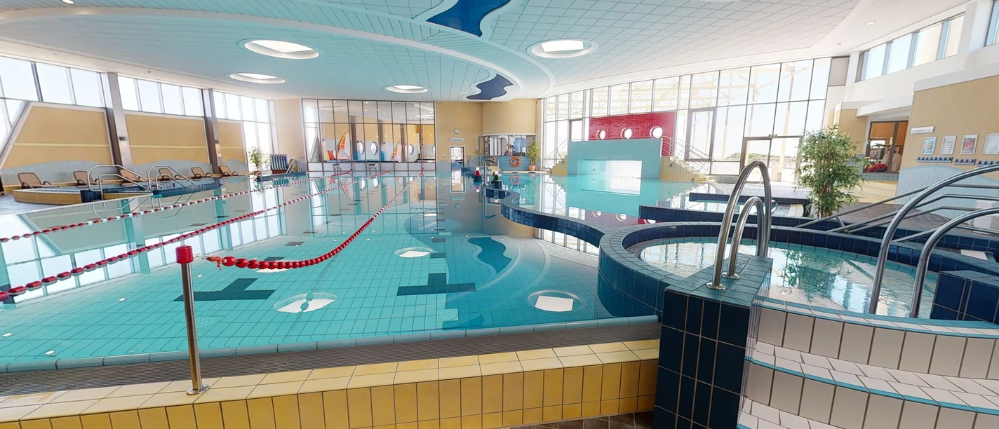 Ein modernes Schwimmbad bietet ein großes Becken mit klar abgegrenzten Schwimmbahnen und eine sanfte, angenehme Beleuchtung, die durch Fenster Tageslicht hereinlässt. Das Design ist einladend und barrierefrei gestaltet.