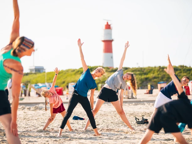 Strandanimation.jpg Eine Gruppe bunt gekleideter Menschen macht Yoga am Strand.