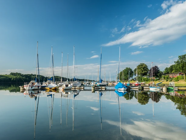 Bevertalsperre Segelboote im ruhigen Wasser, spiegeln sich unter blauem Himmel und weißen Wolken im Hafen wider.