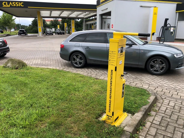 Eine gelbe Lufttankstelle steht auf einem Parkplatz vor einer Tankstelle mit mehreren Autos im Hintergrund.
